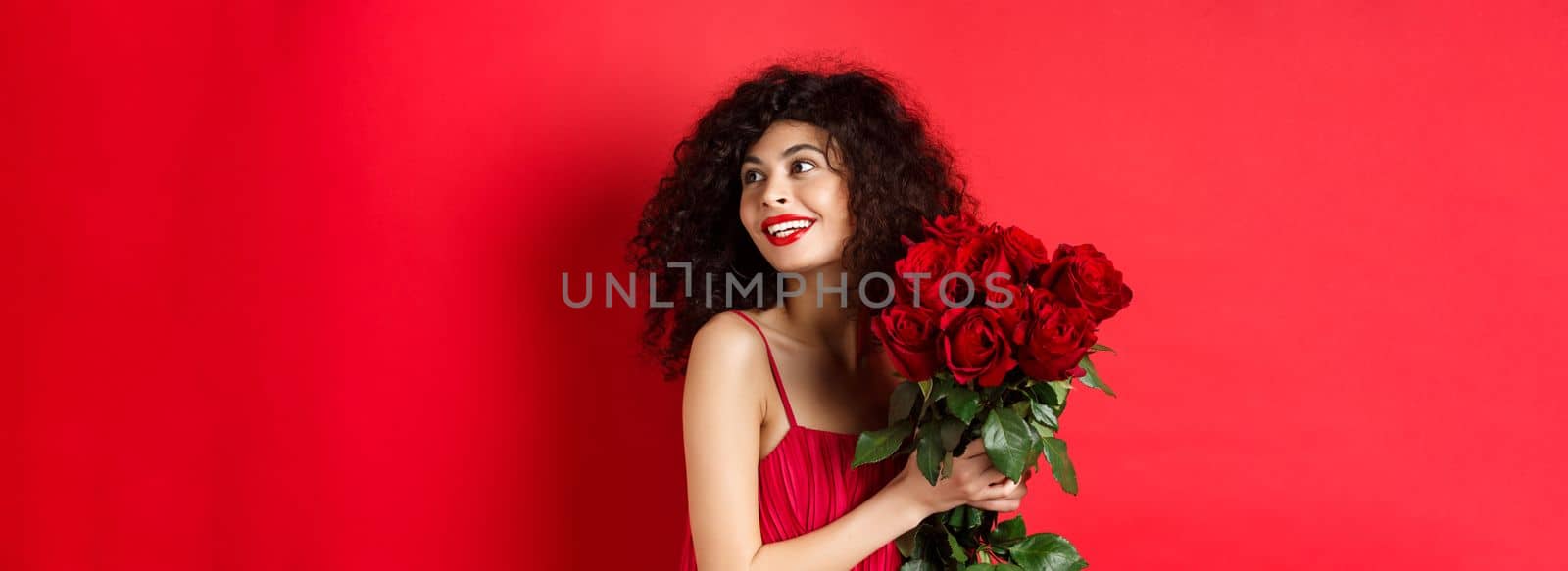 Tender young woman in elegant red dress, holding romantic bouquet of red roses and looking right, smiling dreamy, thinking about lover on Valentines day, studio background by Benzoix
