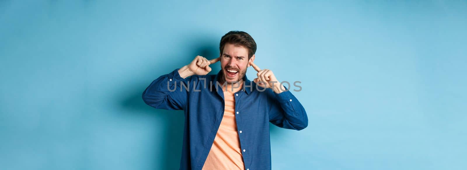 Angry guy shouting with shut ears, block loud music and frowning annoyed, standing on blue background by Benzoix