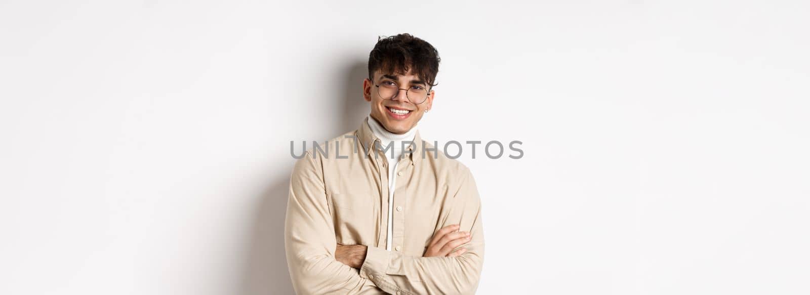 Handsome young man in glasses cross arms on chest, smiling at camera carefree, standing on white background.