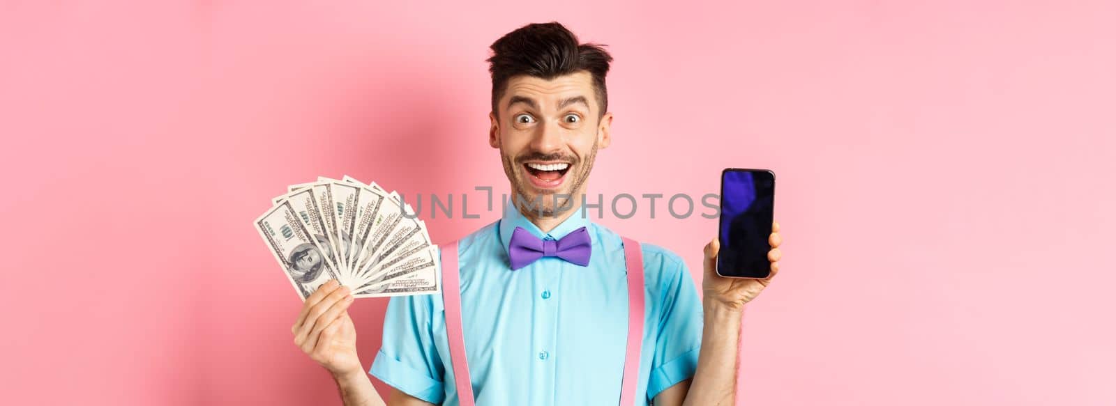 E-commerce and shopping concept. Cheerful guy in bow-tie showing empty smartphone screen and dollars money, smiling amazed at camera, standing on pink background by Benzoix