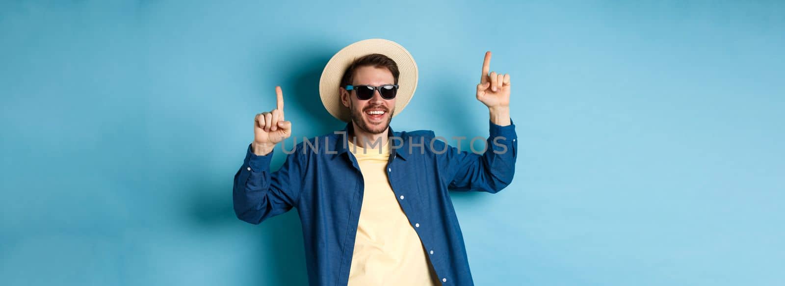 Cheerful caucasian guy in straw hat and sunglasses, dancing and having fun on vacation, standing over blue background. Concept of summer tourism.