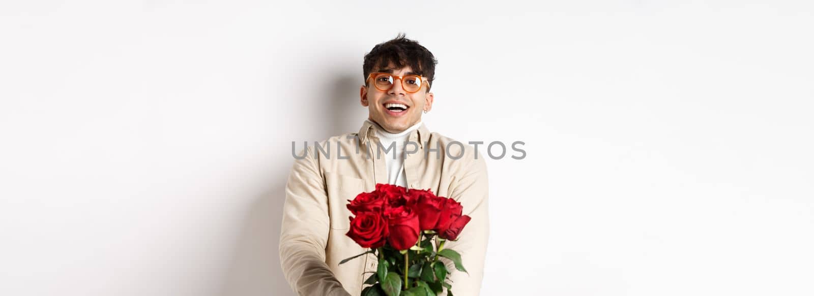 Man in love holding red roses and looking tenderly at camera, staring at lover with happy face, celebrating Valentines day with girlfriend, standing over white background.