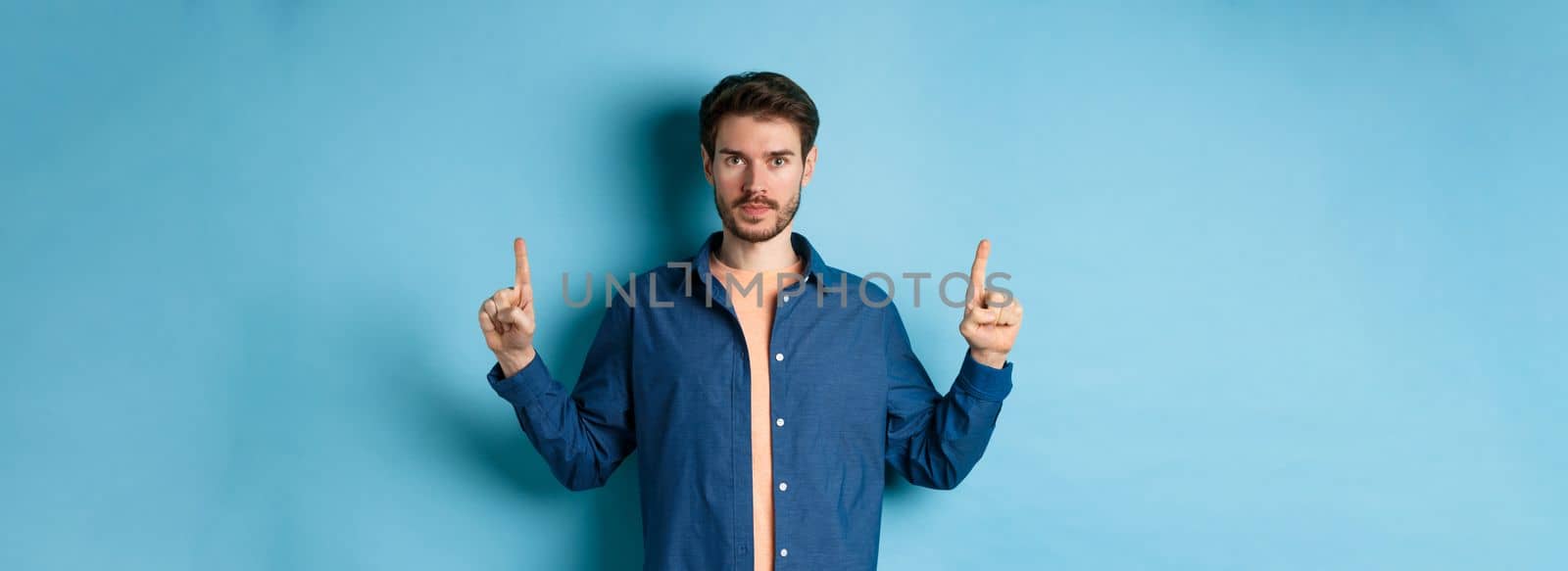 Serious caucasian man with beard pointing fingers up at empty space, showing logo and looking at camera, standing on blue background by Benzoix