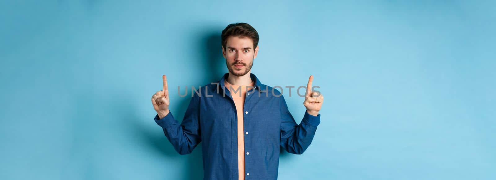 Serious caucasian man with beard pointing fingers up at empty space, showing logo and looking at camera, standing on blue background.