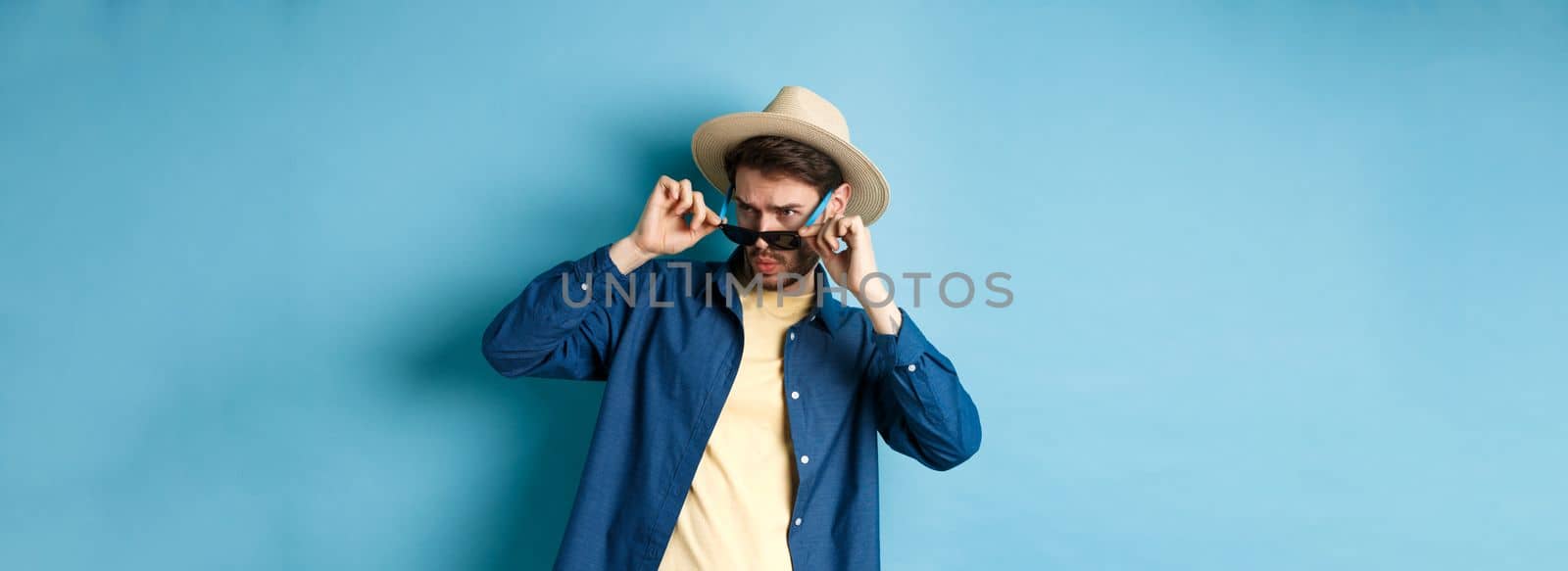 Portrait of guy on vacation staring aside with shocked and cringe face, take-off sunglasses and look at empty space on blue background by Benzoix