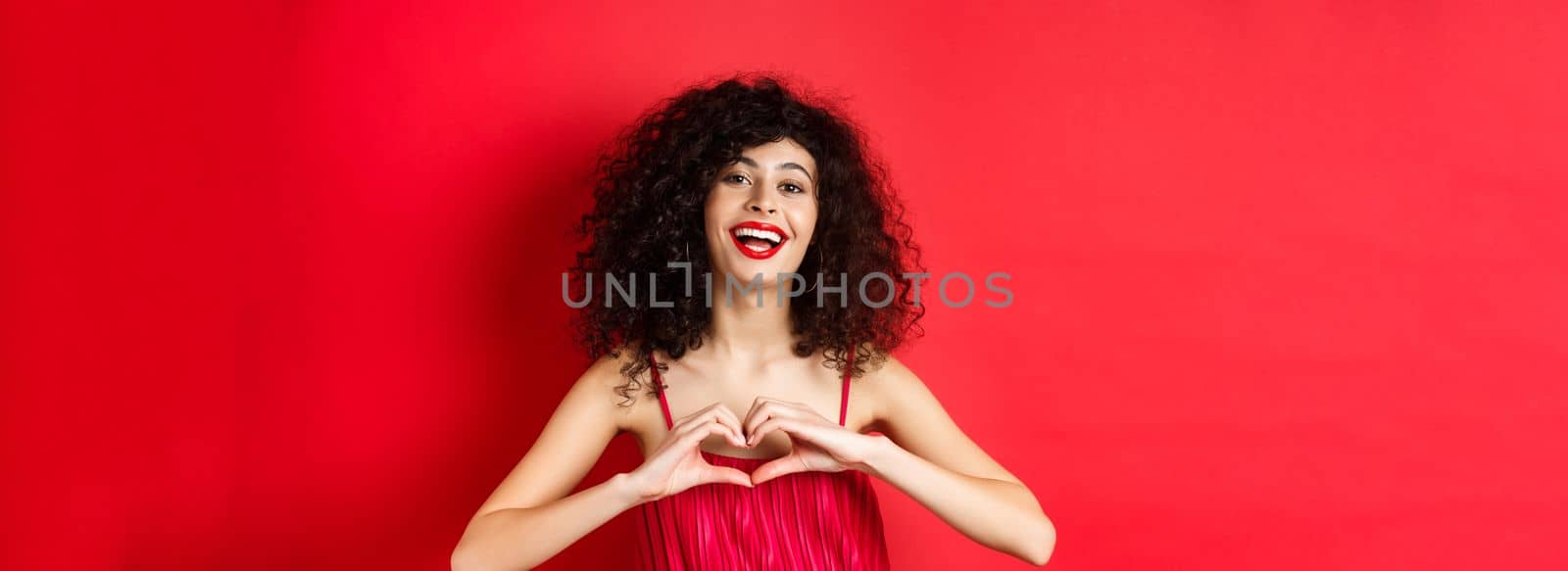 Beautiful woman with curly hair, red dress, showing heart symbol and smiling happy, studio background.