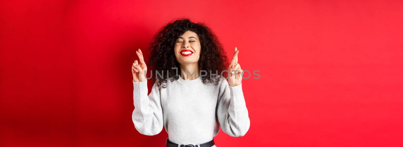 Hopeful and positive woman with red lips and curly hair, cross fingers for good luck and making wish, praying for dream come true, smiling excited, red background.