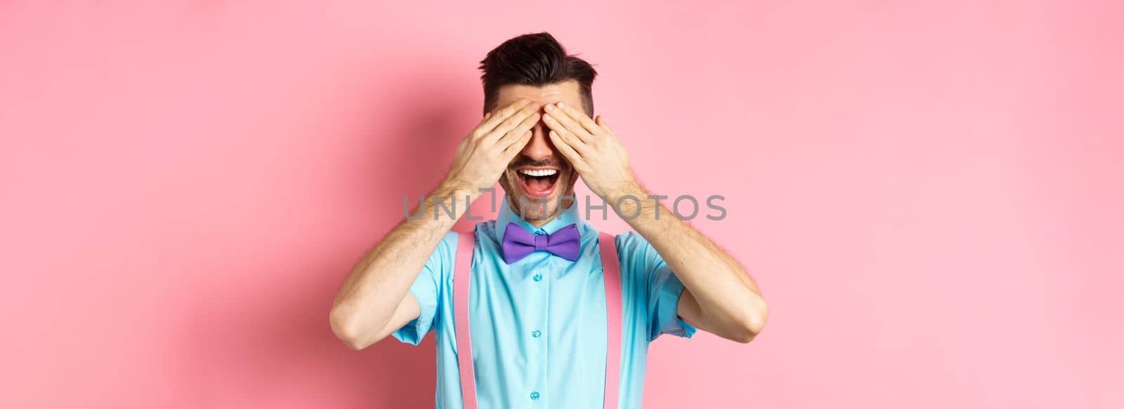 Cheerful young man celebrating, waiting for surprise with closed eyes and happy smile, standing joyful on pink background.