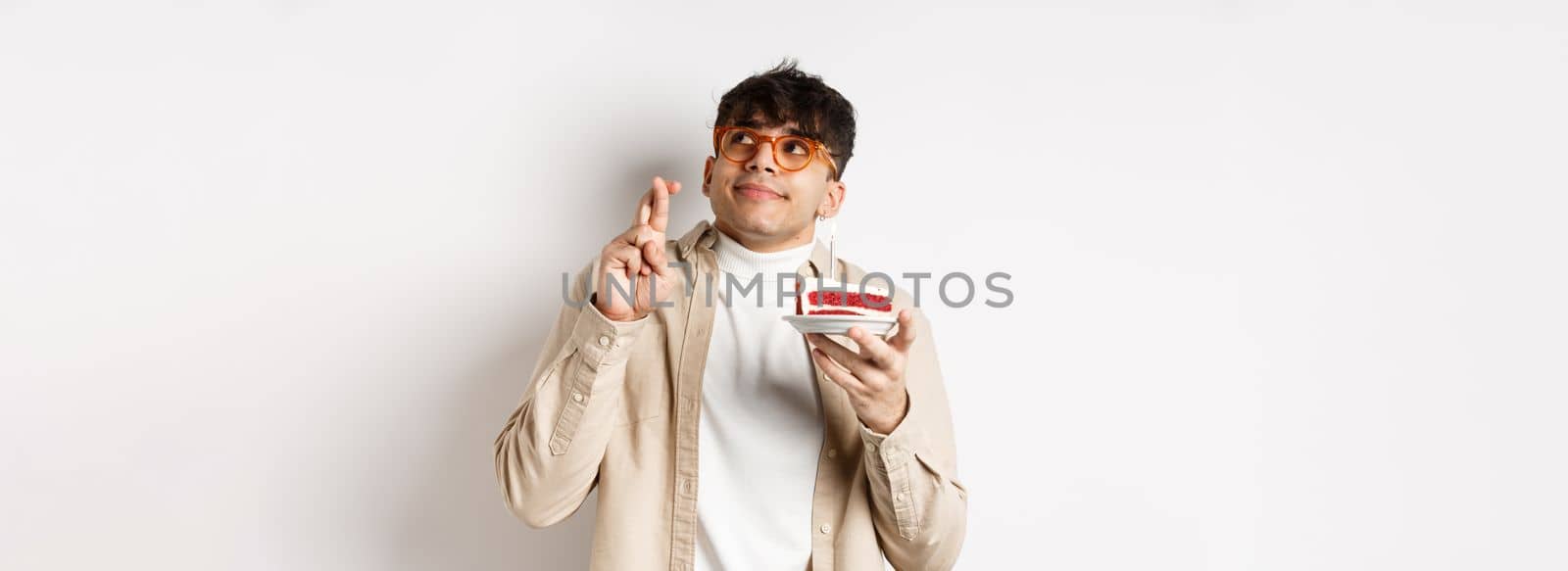 Cute hopeful guy in glasses making birthday wish, holding cake and fingers crossed, looking up and praying, hope dream come true, standing on white background by Benzoix
