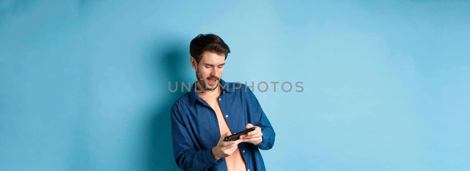 Young guy playing video game on mobile phone, tilt body and holding smartphone horizontally, standing on blue background.