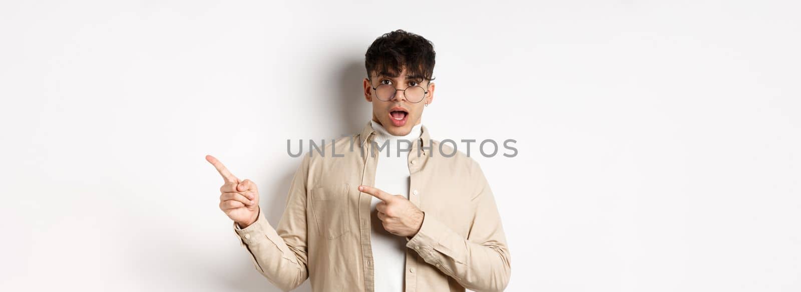 Shocked young man in glasses pointing fingers right at empty space, asking about advertisement, standing on white background by Benzoix