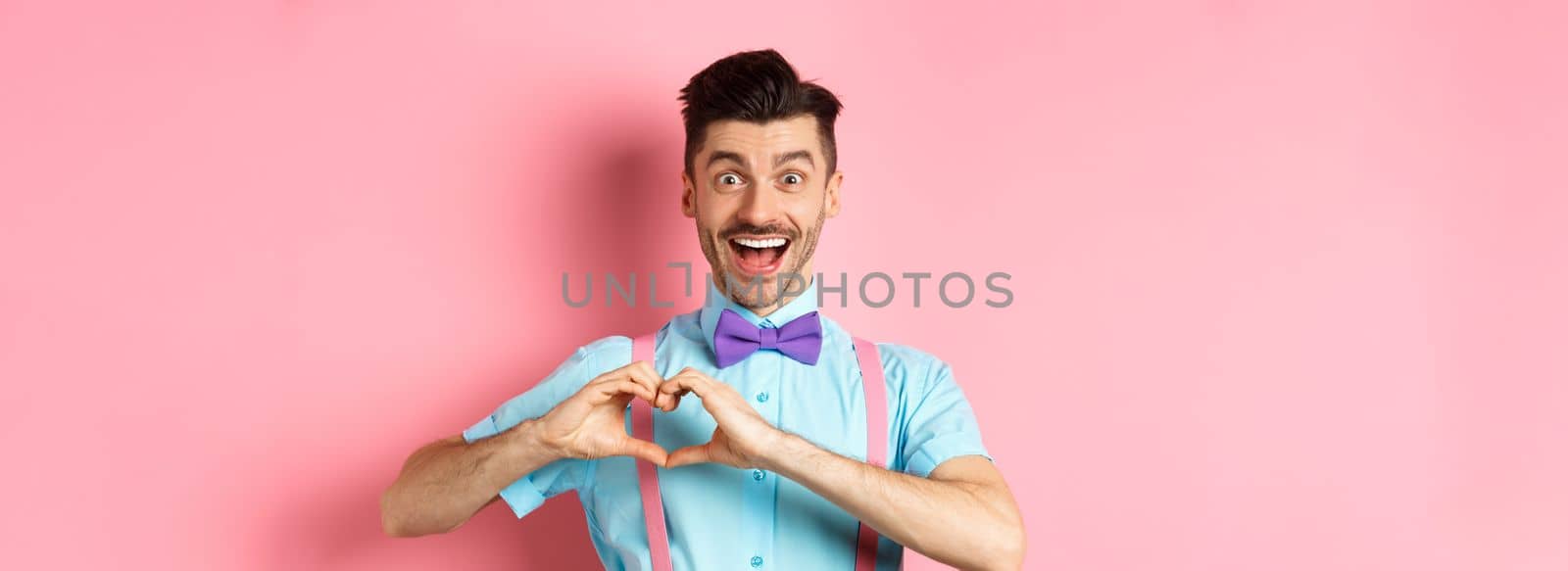 Excited smiling man showing pounding heart and looking with love, standing over romantic pink background. Valentines day concept.