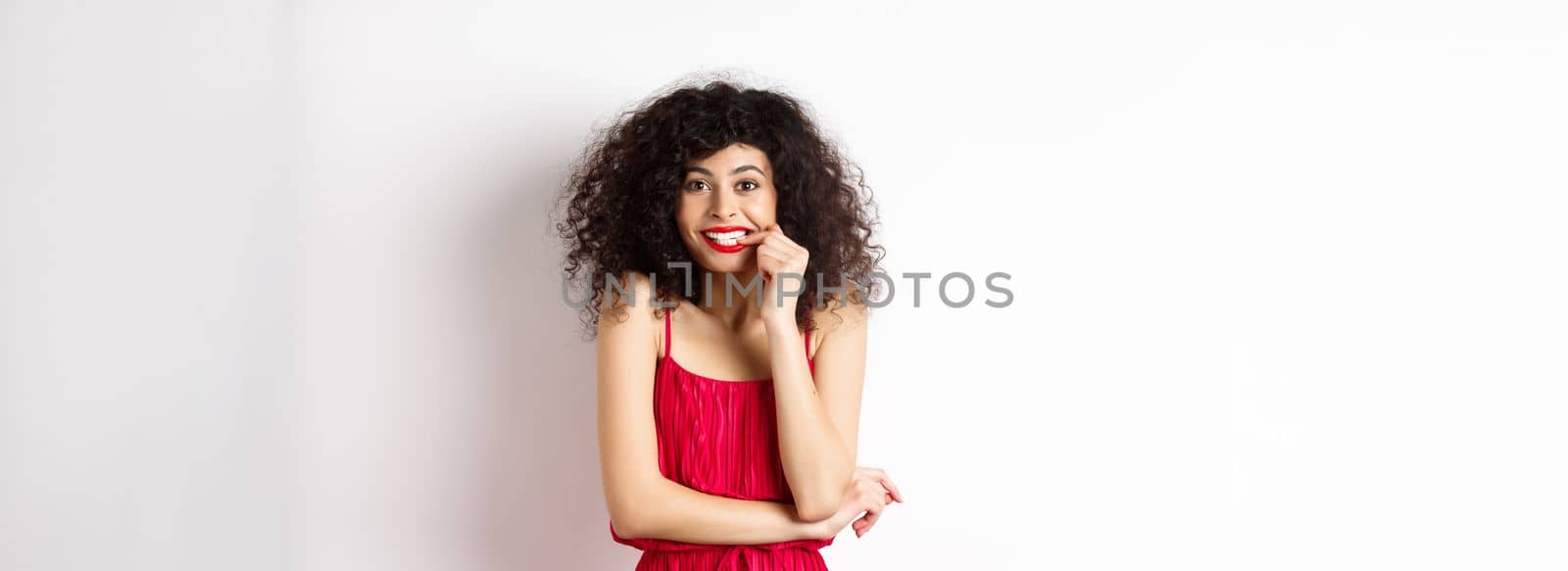 Intrigued young woman with curly hair, wearing elegant red dress and lipstick, biting fingernails and looking interested, standing over white background.