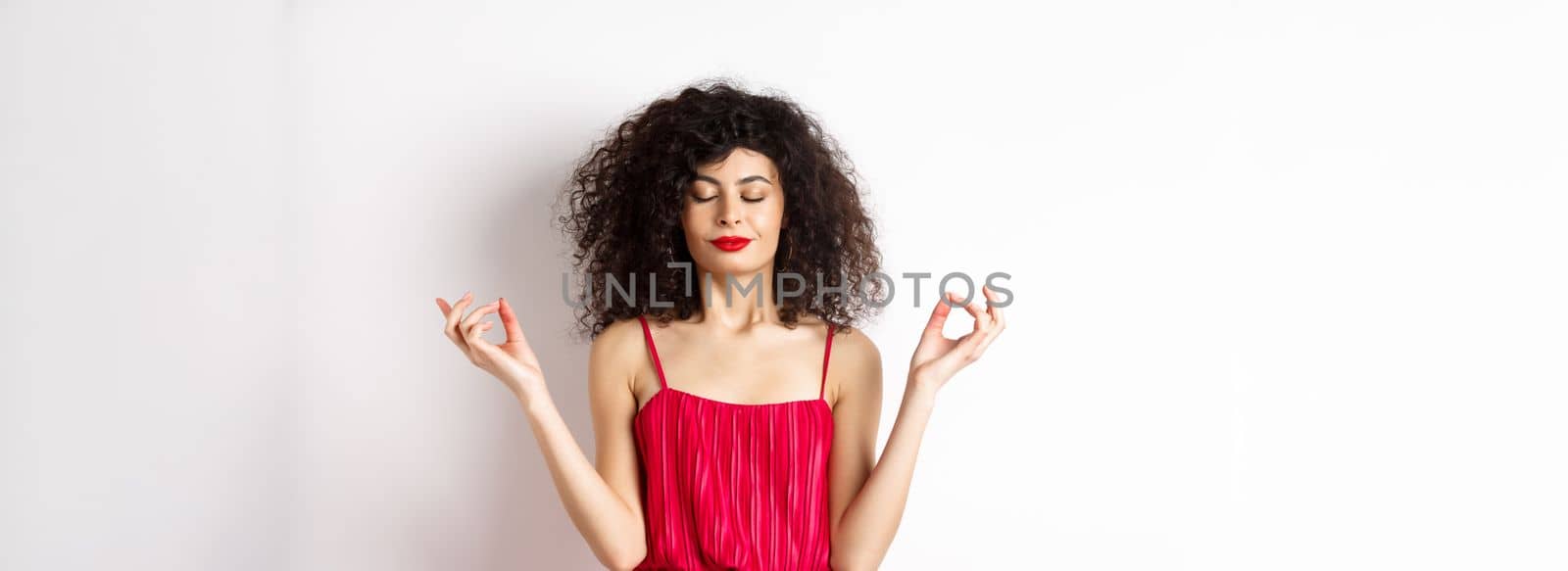 Calm and relaxed smiling woman in red dress, close eyes and meditating, practice yoga in zen pose, standing on white background by Benzoix