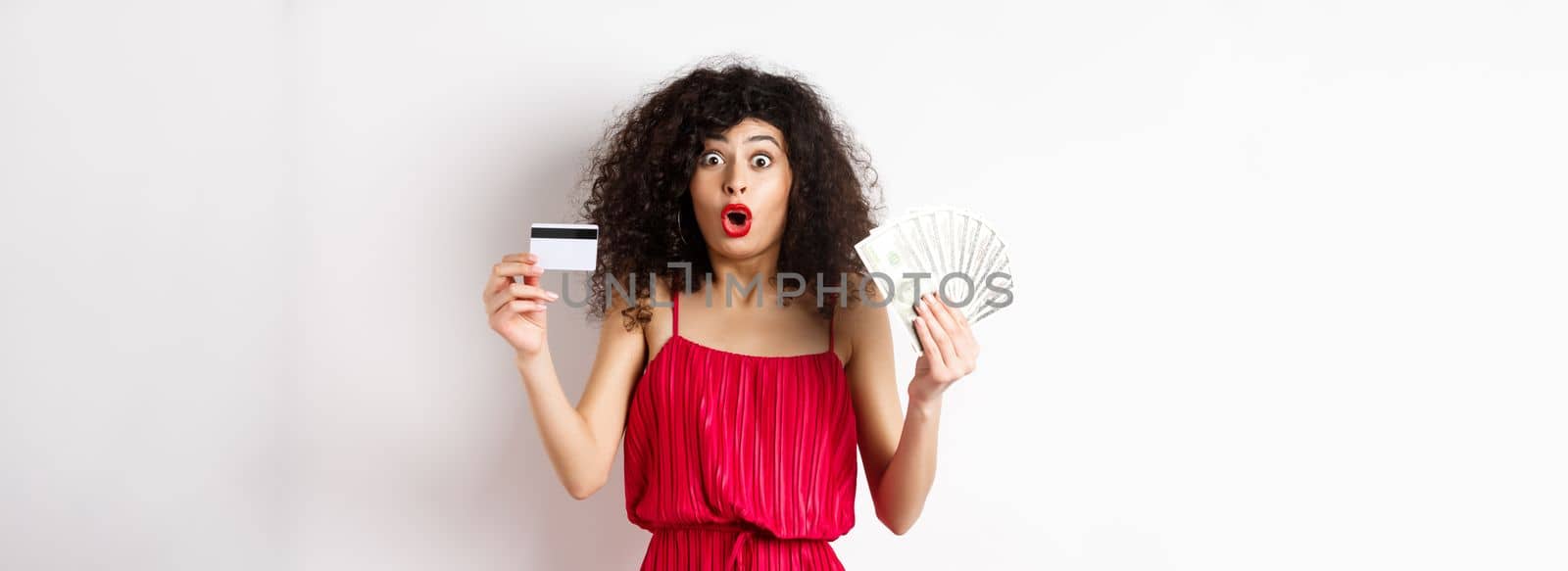 Impressed lady with curly hair and trendy red dress, saying wow, showing plastic credit card with cash, standing on white background.
