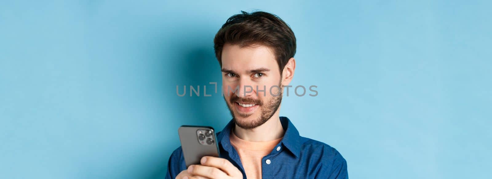 Close-up of attractive caucasian man holding mobile phone and smiling at camera, standing in casual outfit on blue background.