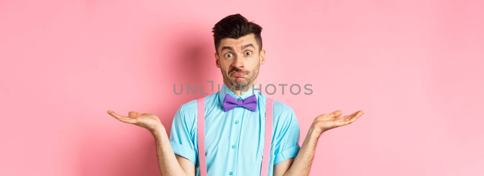 Image of confused guy in bow-tie and suspenders know nothing, shrugging shoulders and looking clueless, standing over pink background by Benzoix