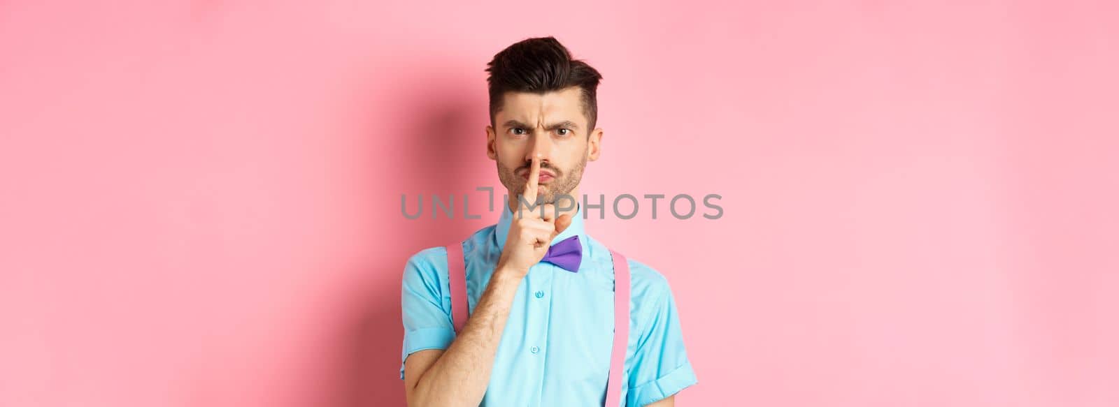 Angry and grumpy man in funny bow-tie shushing at camera, frowning and demand silence, tell to be quiet, scolding someone loud, standing on pink background.