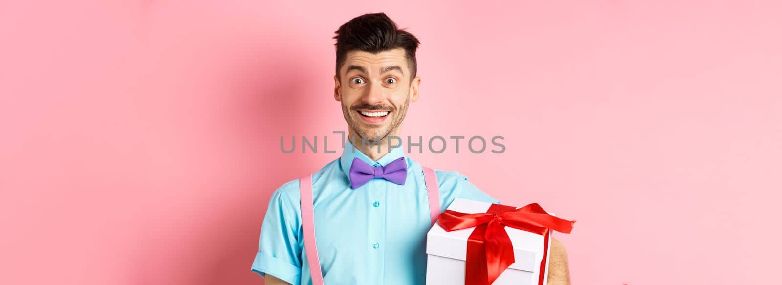 Holidays and celebration concept. Cheerful guy in bow-tie and suspenders bring gift box to party, holding present and smiling, standing over pink background by Benzoix