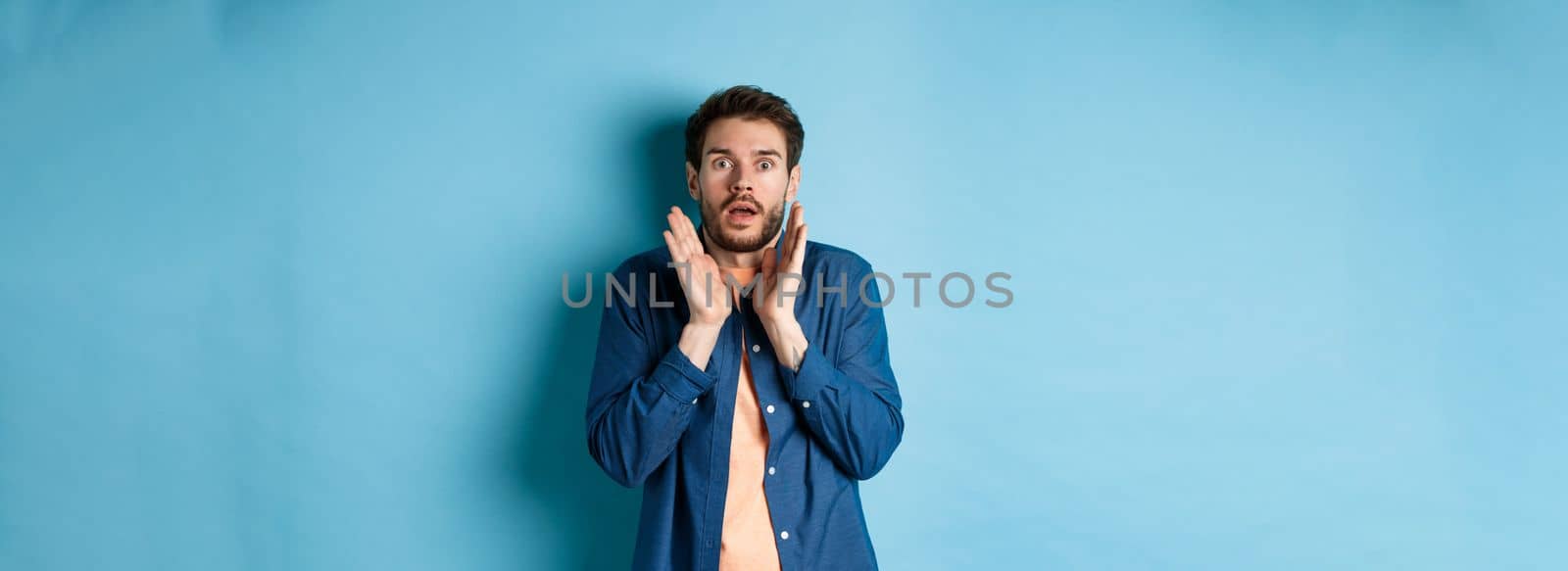 Shocked and scared young man standing in stupor, raising hands up and gasping speechless, standing on blue background by Benzoix