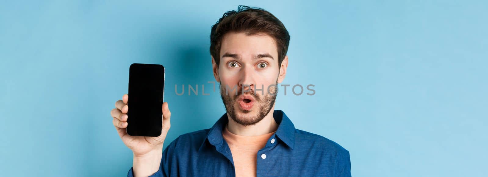 Close up of impressed caucasian man say wow, showing empty smartphone screen, standing on blue background by Benzoix