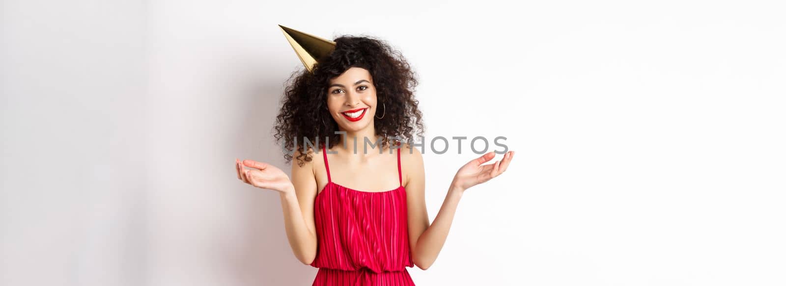 Beautiful caucasian woman in red dress, celebrating holiday, wearing party hat and smiling, standing on white background by Benzoix