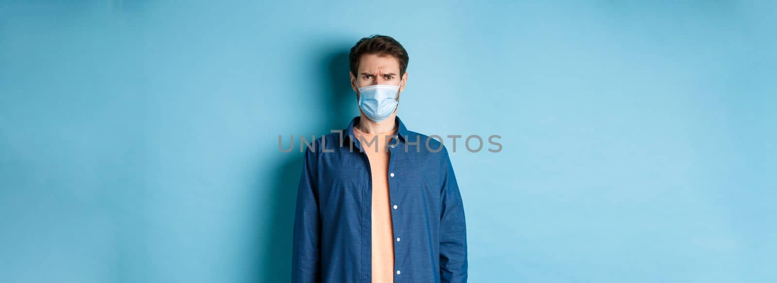 Covid-19 and healthcare concept. Disappointed guy in medical mask frowning, looking confused, standing on blue background.