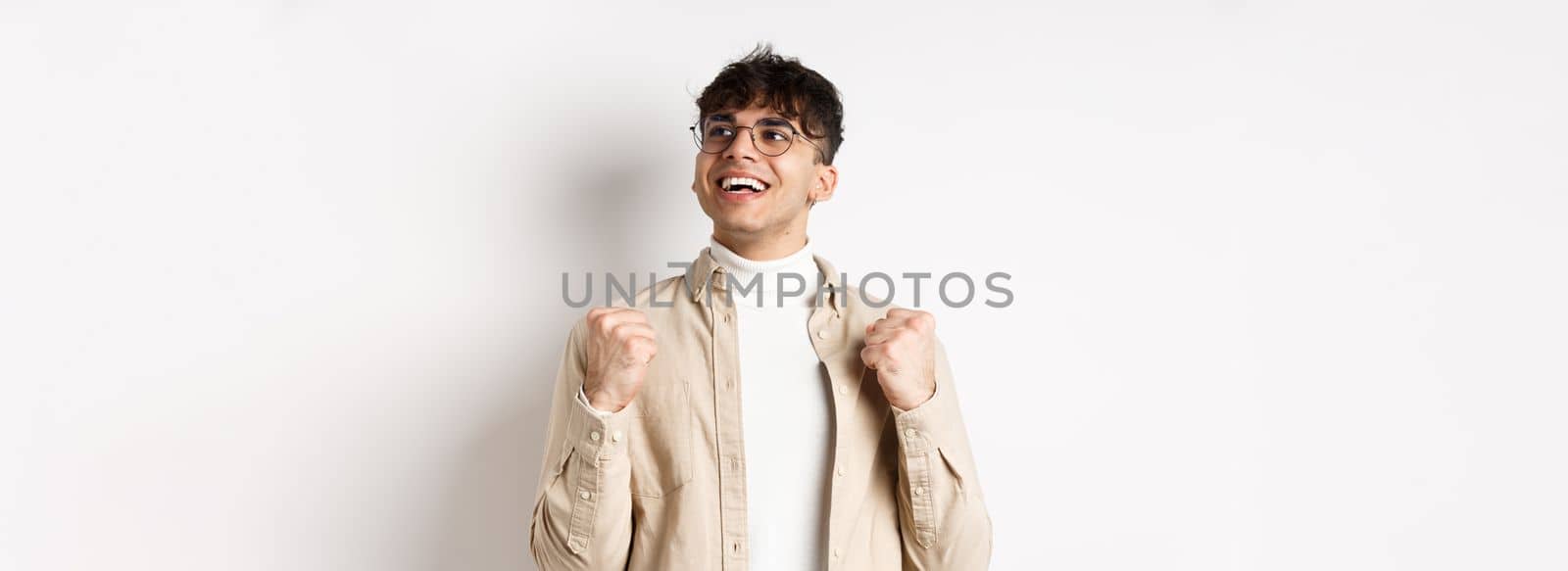 Lucky young man say yes with relieved face, smiling and making fist pump, triumphing of success, winning prize, standing on white background after achieving goal by Benzoix