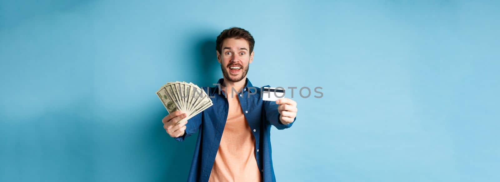 Cheerful caucasian guy extending hands with dollars and plastic credit card, smiling happy at camera, standing on blue background by Benzoix