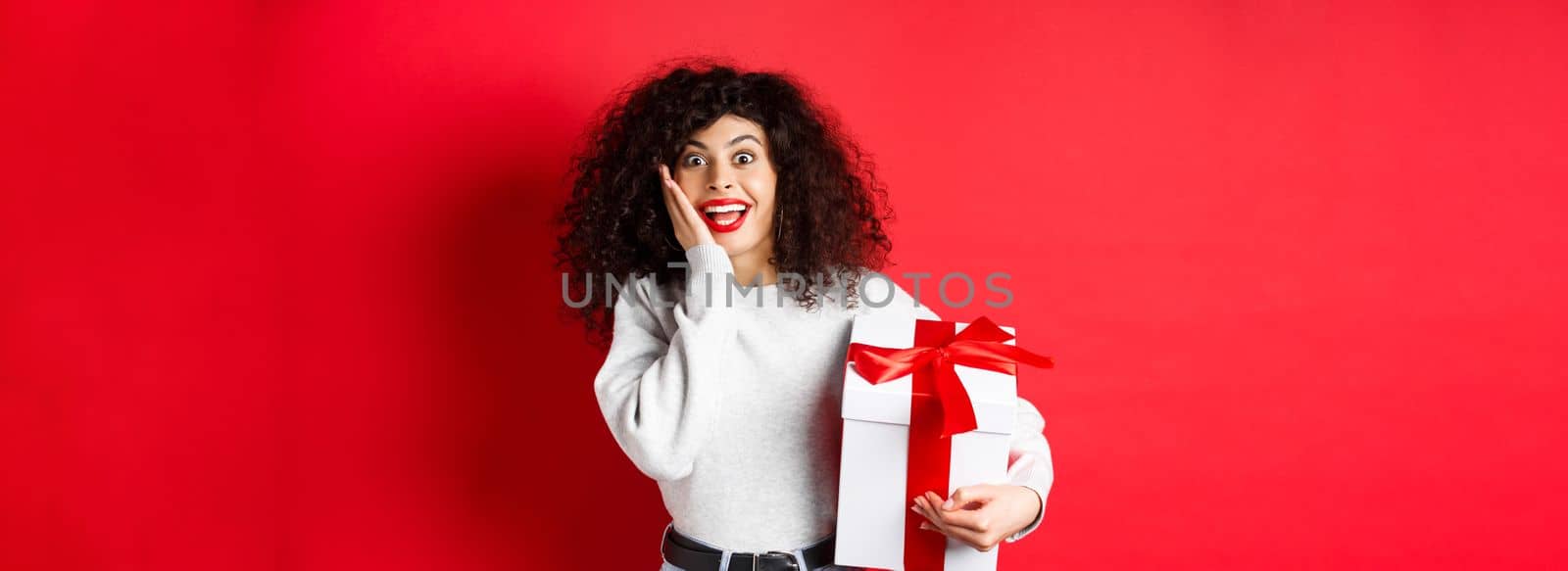 Surprised pretty girl receive Valentines day romantic gift, holding present box and looking with disbelief and happiness at camera, standing on red background.