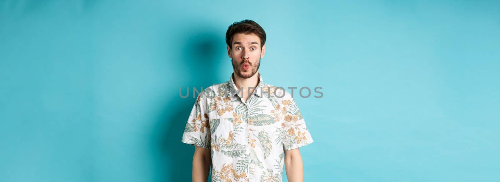 Summer holiday. Surprised tourist say wow and staring at camera, checking out awesome promo, standing in hawaiian shirt on blue background.