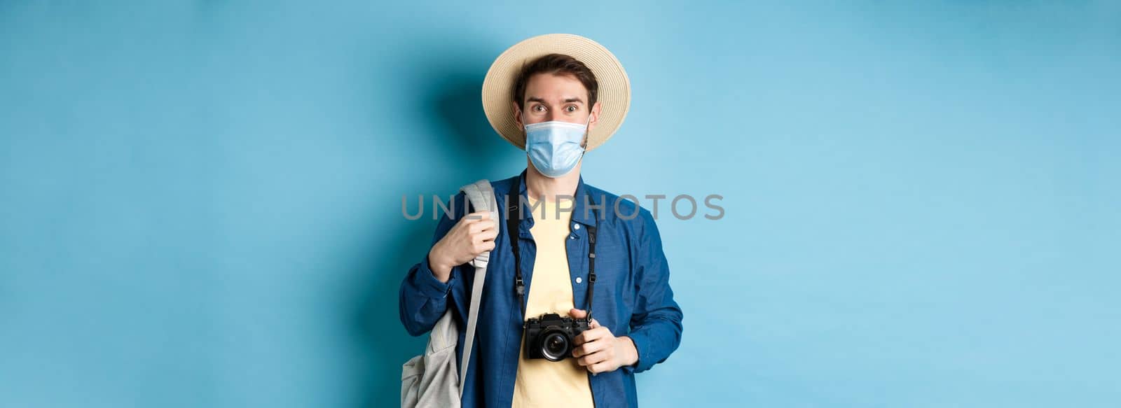 Covid-19 and travelling concept. Young guy tourist in medical mask and summer hat travel abroad during coronavirus pandemic, taking pictures on vacation, blue background by Benzoix