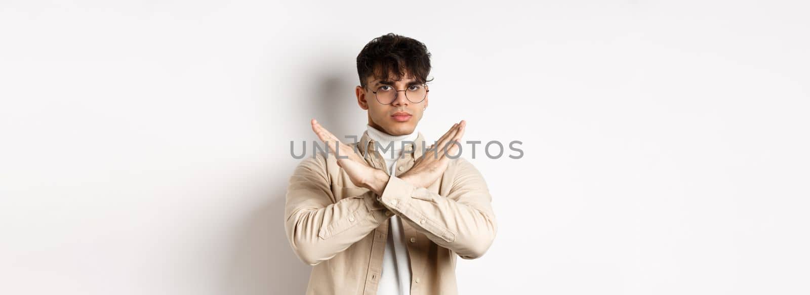 Image of serious young guy in glasses say no, showing cross gesture, make stop sign to disagree or prohibit something, standing on white background.