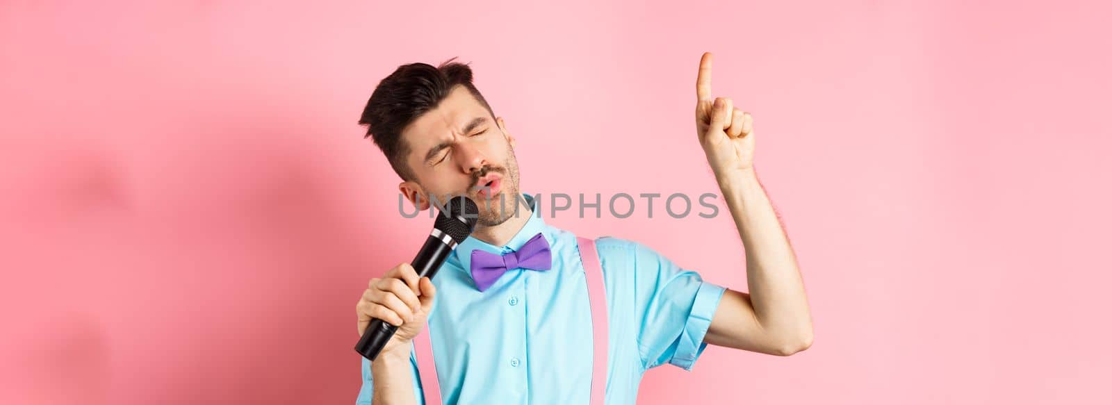 Party and festive events concept. Funny guy singing in microphone, raising finger up as reaching high note in song, standing on pink background.