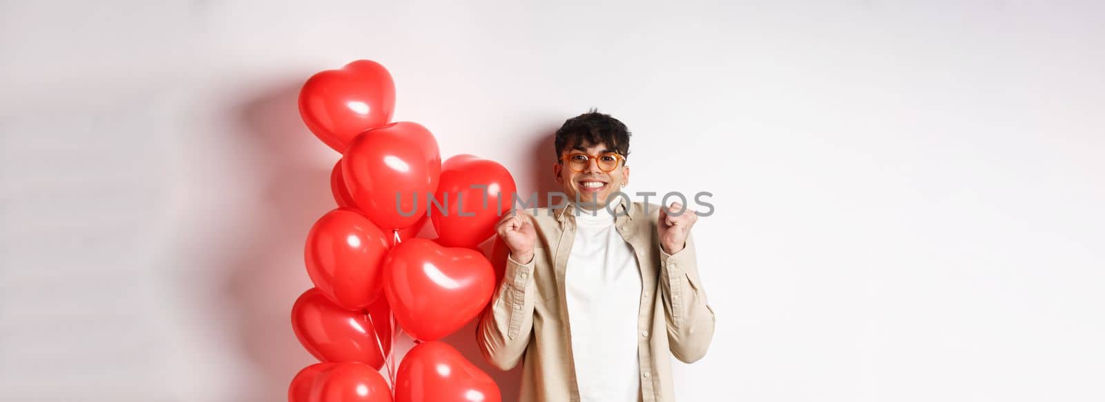 Valentines day. Excited smiling man eager to go on date, celebrating with lover, standing near red hears balloons, white background.