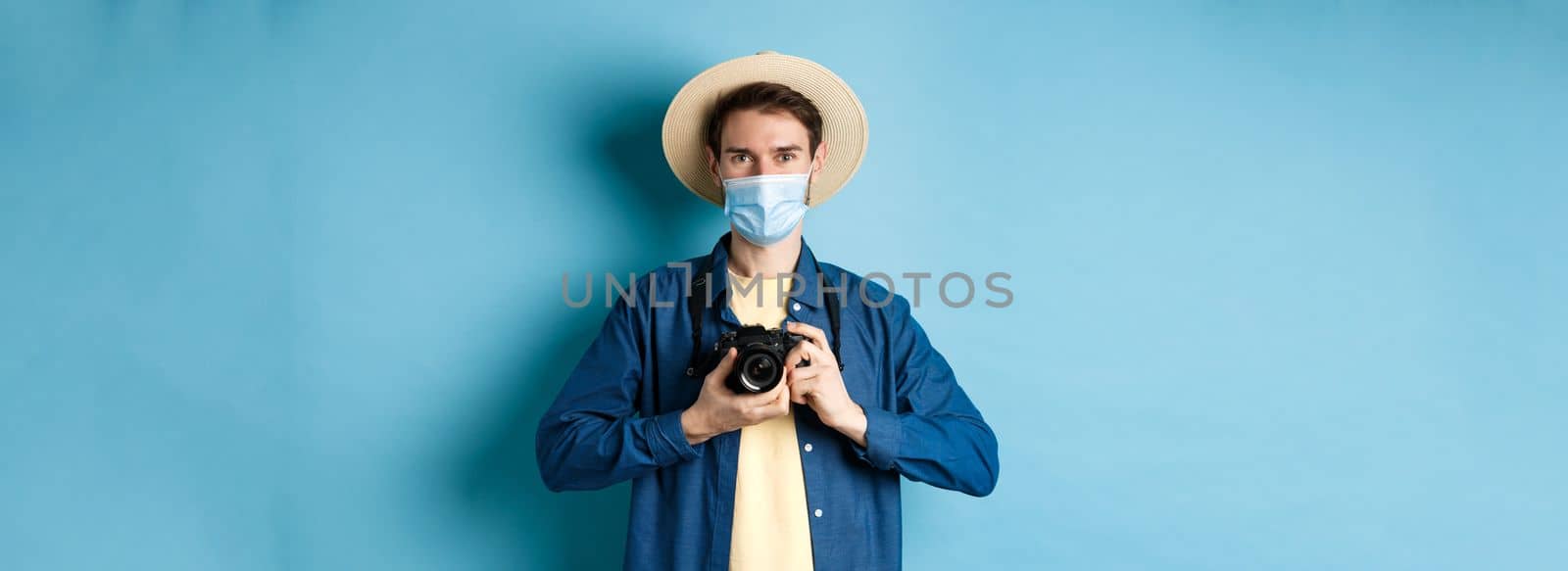 Covid-19, pandemic and travel concept. Happy guy on vacation taking pictures with camera, wearing medical mask and summer hat, blue background by Benzoix