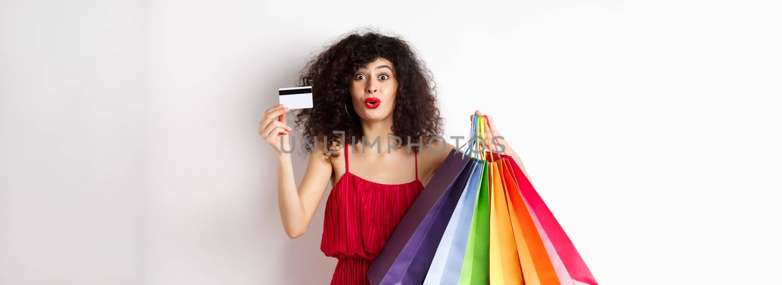 Excited woman in red dress, holding shopping bags and showing plastic credit card, shop with discounts, white background.