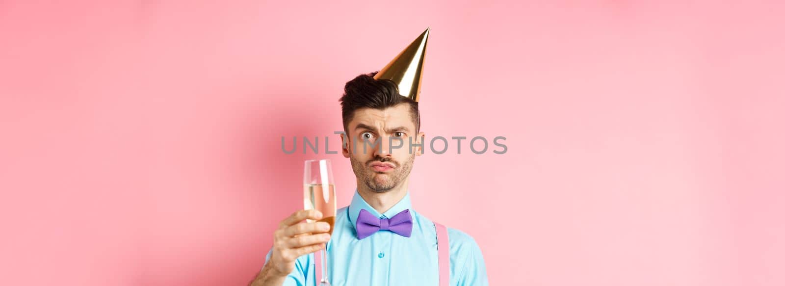 Holidays and celebration concept. Troubled young man in party hat, frowning with doubtful face, raising glass of champagne perplexed, standing on pink background by Benzoix