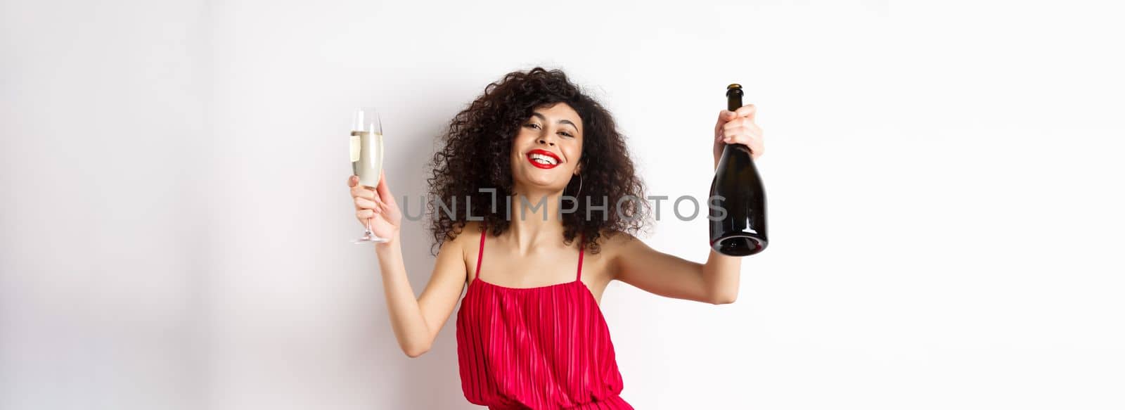 Happy party girl in red dress, dancing with bottle of champagne and glass, drinking and having fun, celebrating holiday, standing on white background.