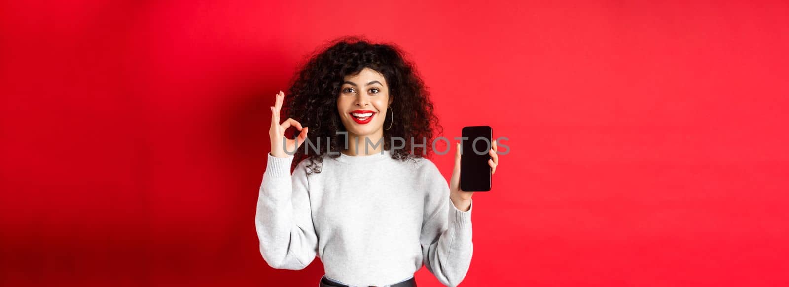 Cheerful european woman with curly hair, showing empty mobile phone screen and okay gesture, smiling satisfied, praise good app or promotion, red background.