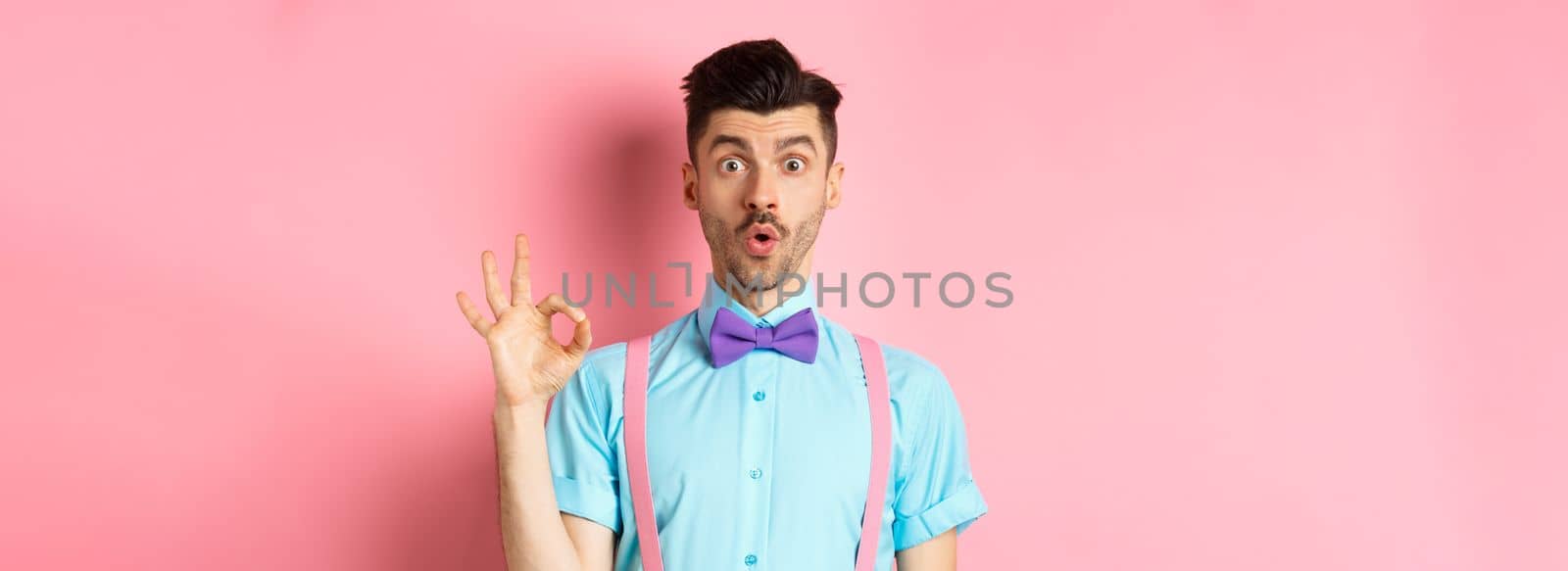 Impressed guy in bow-tie and suspenders saying wow, showing okay gesture with amazement, praise awesome thing, standing over pink background.