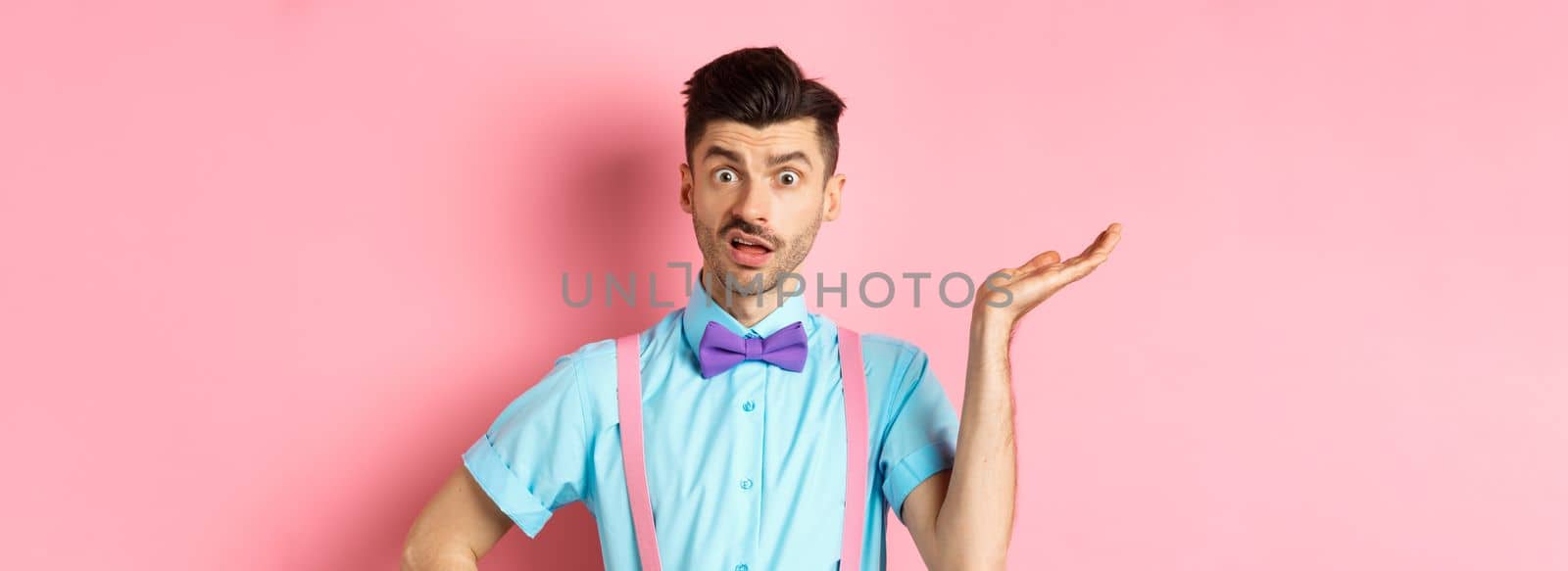 Confused guy cant understand what happening, looking with wtf face, raising hand and staring at camera shocked, standing over pink background by Benzoix