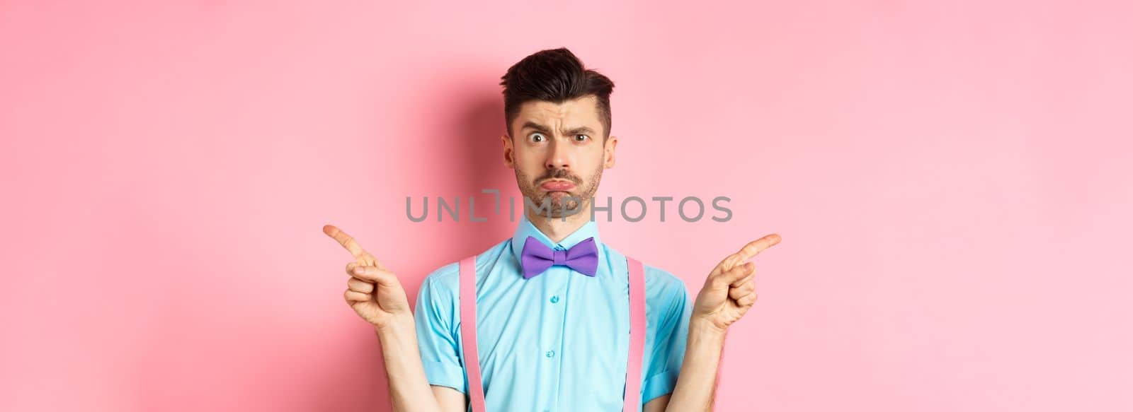 Indecisive young man pucker lips and frowning, pointing fingers sideways at two things, cant decide, standing puzzled over pink background by Benzoix