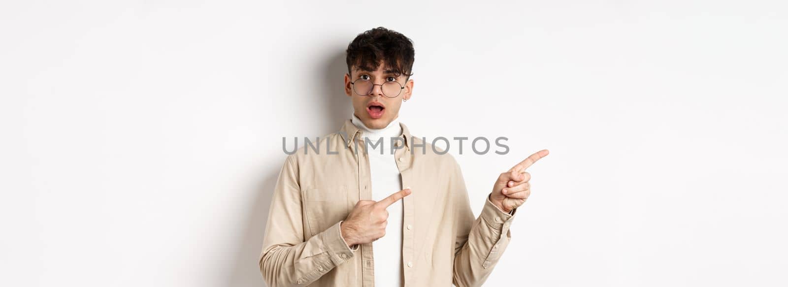 Portrait of young man in glasses drop jaw impressed, pointing fingers left at empty space with amazed face, standing on white background.