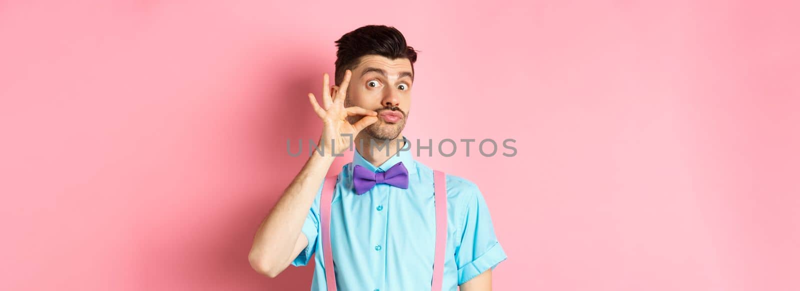 Funny young man touching his french moustache and pucker lips, looking silly at camera, standing in bow-tie and suspenders on pink background by Benzoix