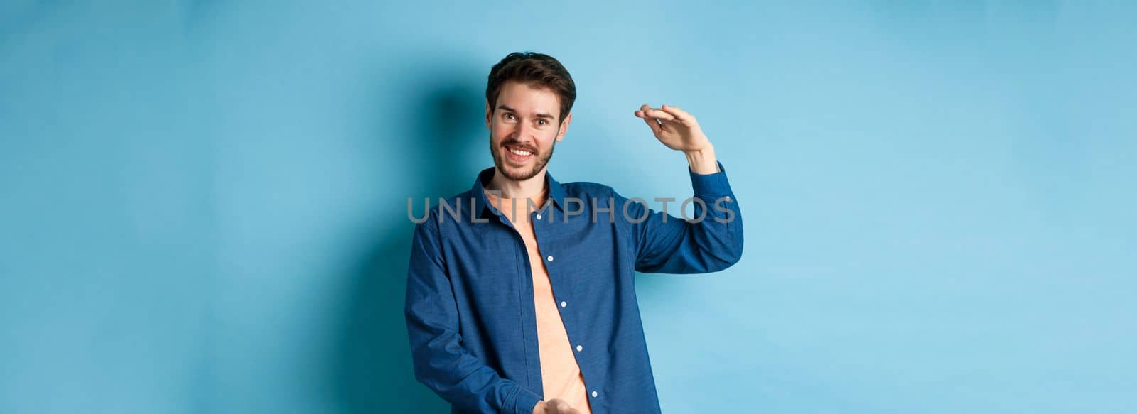 Handsome caucasian guy showing something big, shaping large size object with hands and smiling, standing on blue background.