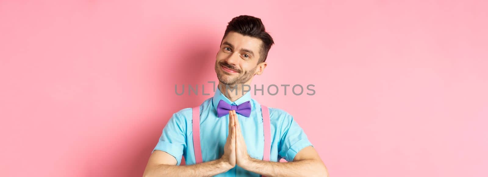 Smiling young guy in bow-tie say thank you, looking with gratitude and holding hands in namaste gesture, standing over pink background by Benzoix