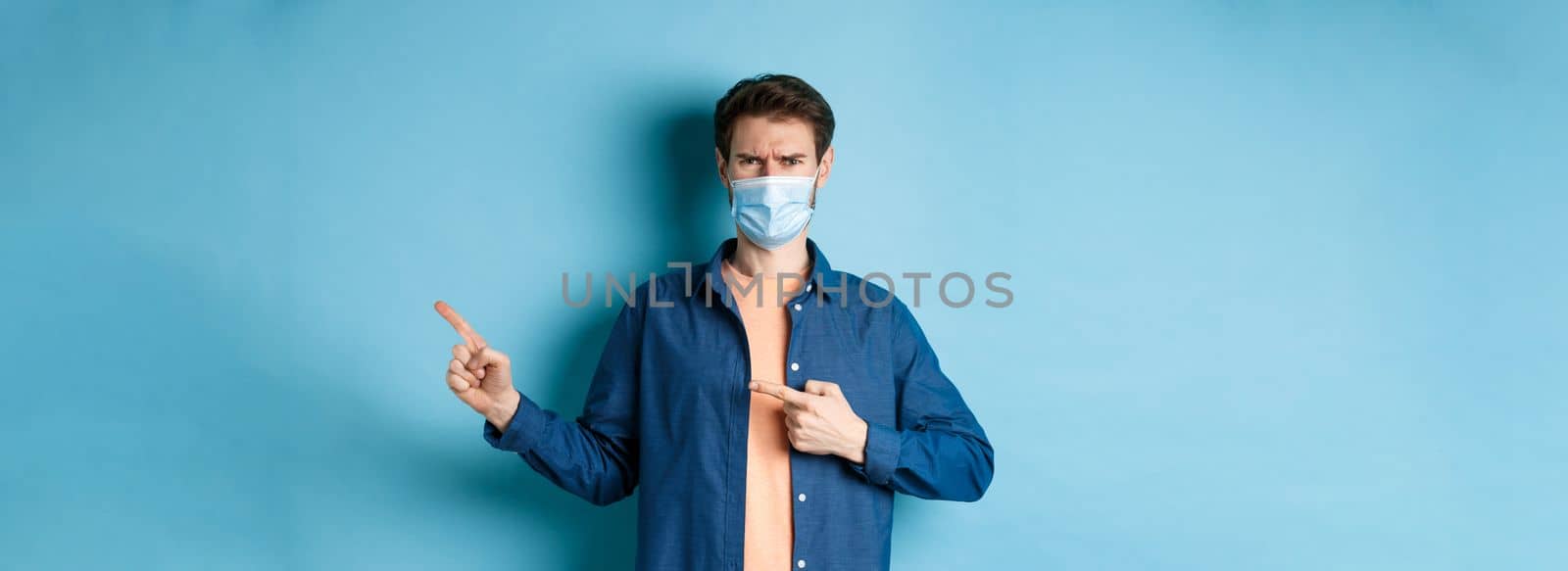 Covid-19 and pandemic concept. Angry man in medical mask frowning, pointig fingers at upper right corner empty space, complaining, standing on blue background by Benzoix