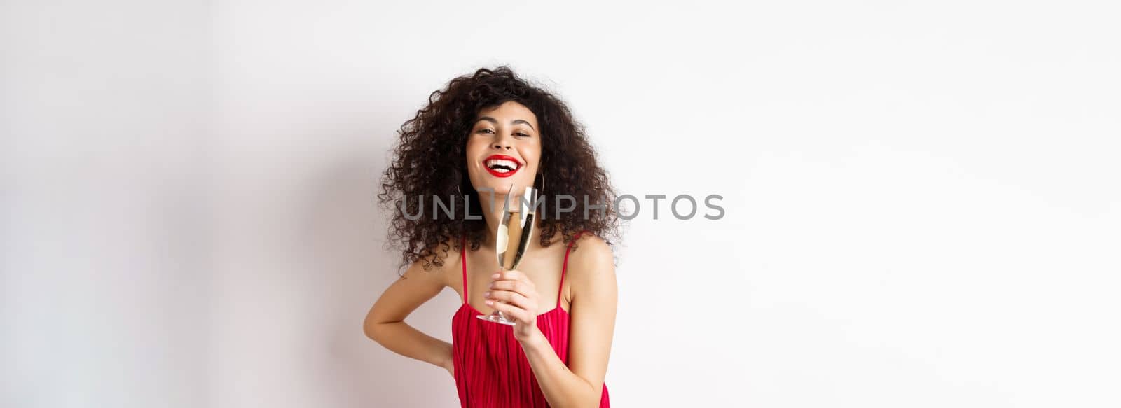 Cheerful elegant woman with dark curly hair, wearing party dress, celebrating anniversary on valentines day, drinking champagne from glass and smiling at camera, white background.