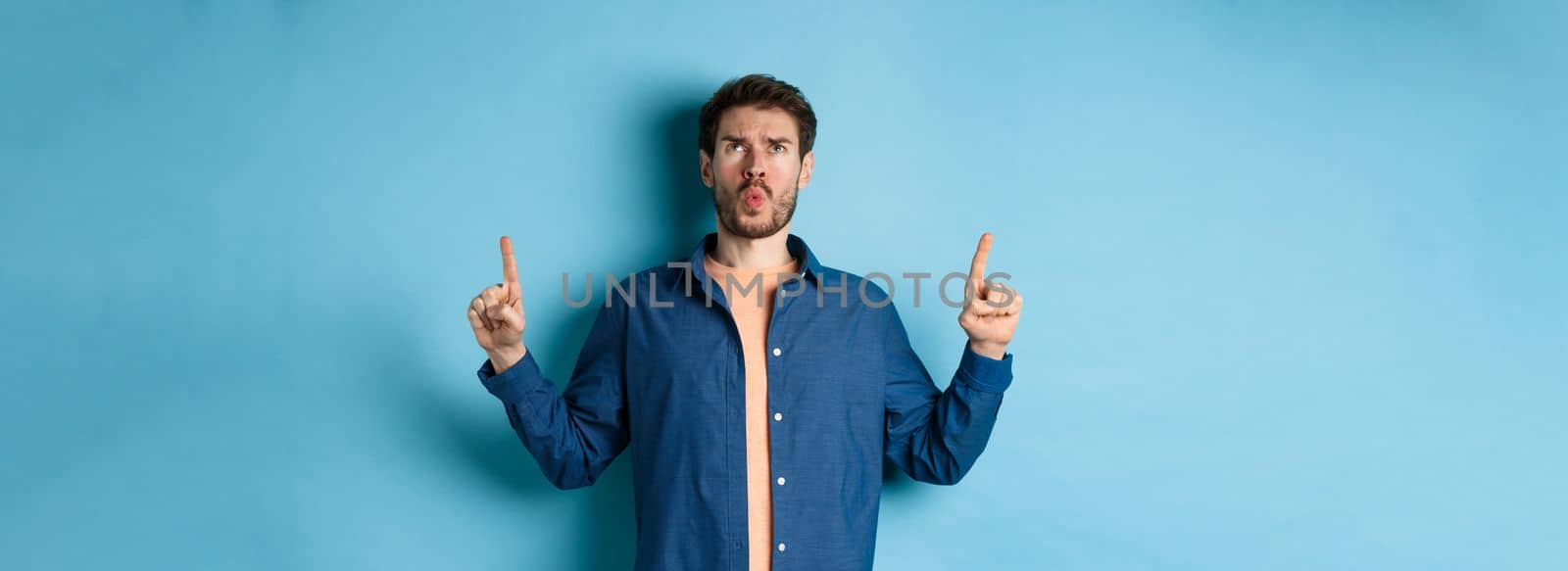 Confused young man frowning, pointing fingers up and looking displeased, standing on blue background. Copy space